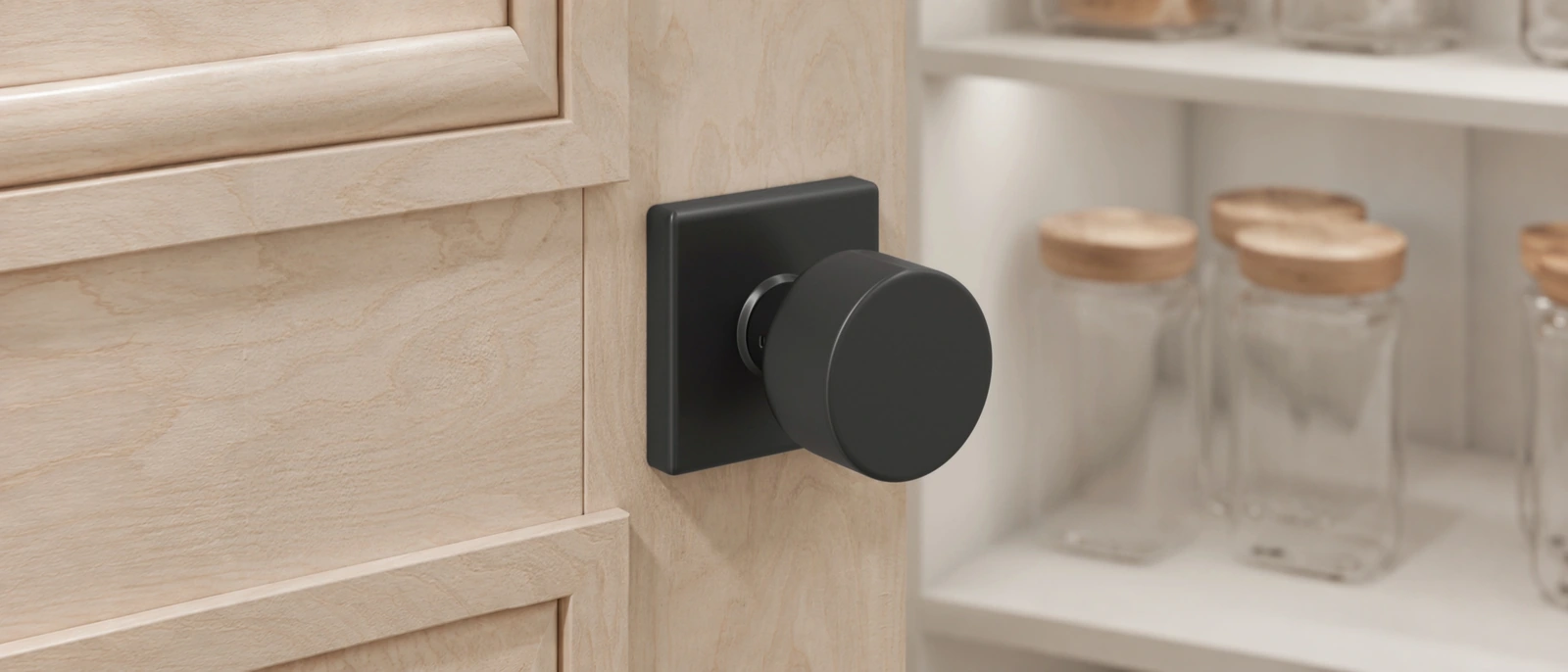Matte-black round knob on a square backplate against pale wood, with softly blurred pantry jars behind